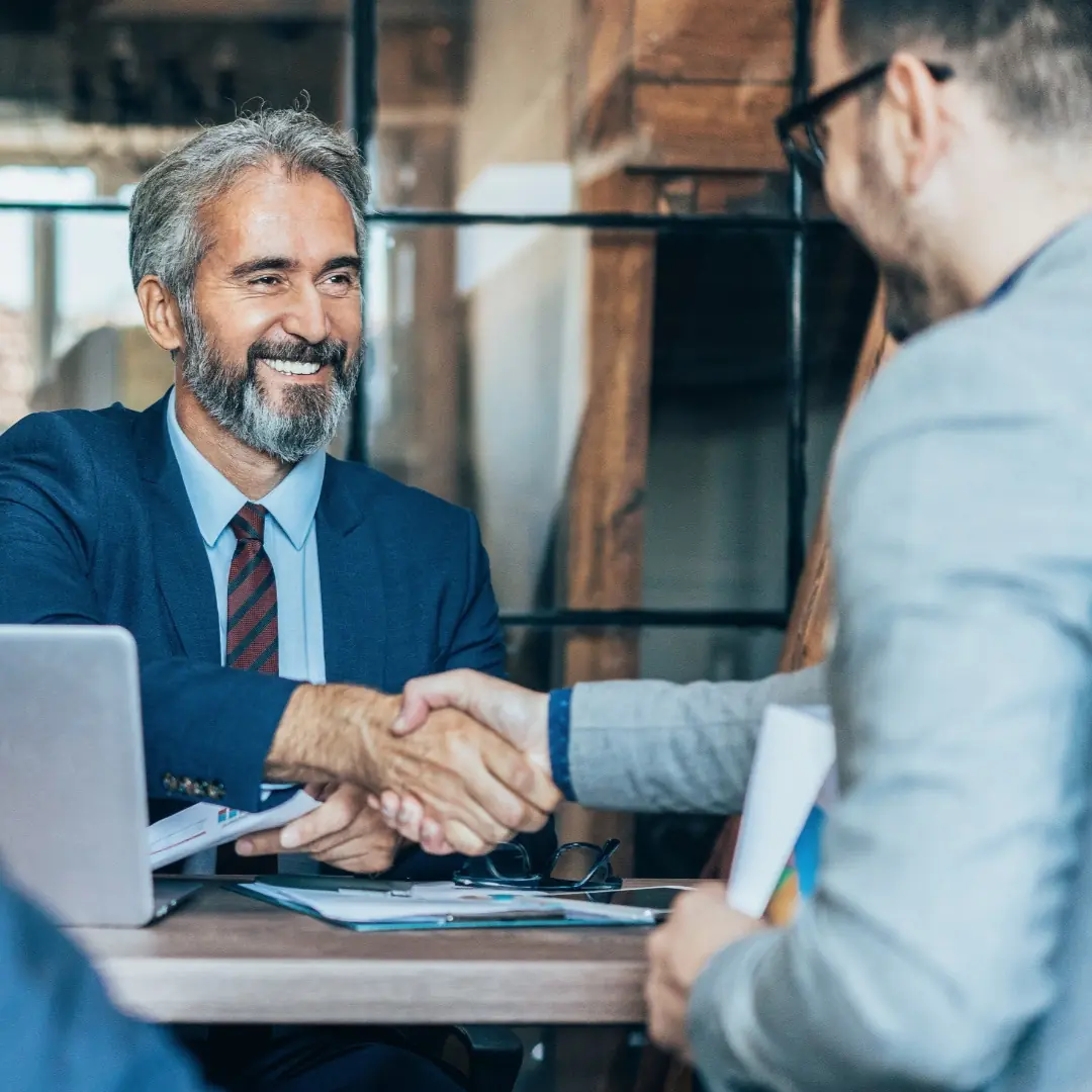 Kaksi miestä kättelee pöydän ääressä sopimuksen merkiksi